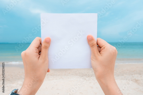 Woman hands holding white paper with sea beach and blue sky background.