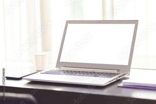 Laptop on wooden table near the window at the office with white screen. White notebook on the table with a cup of coffee in the building with clipping path on white screen.