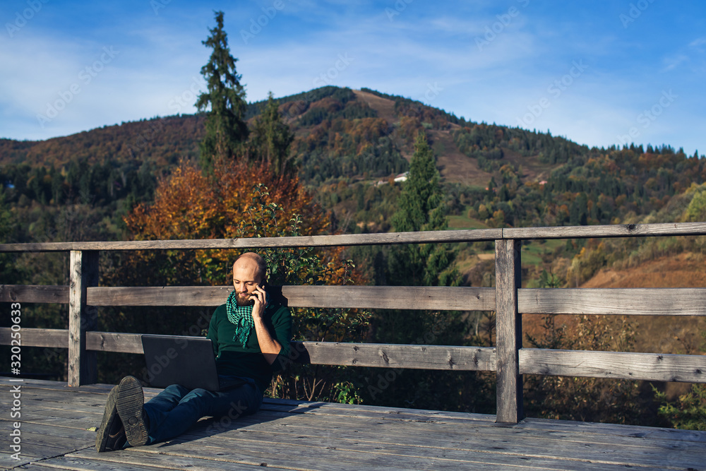 Bearded businessman sitting on terrace in mountains with laptop and talking on phone. Bearded businessman sitting on terrace in mountains with laptop and talking on phone.