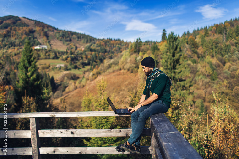 bearded businessman sits on railing and works on laptop. Freelancer hipster on vacation in mountains. bearded businessman sits on railing and works on laptop. Freelancer hipster on vacation in mountains.