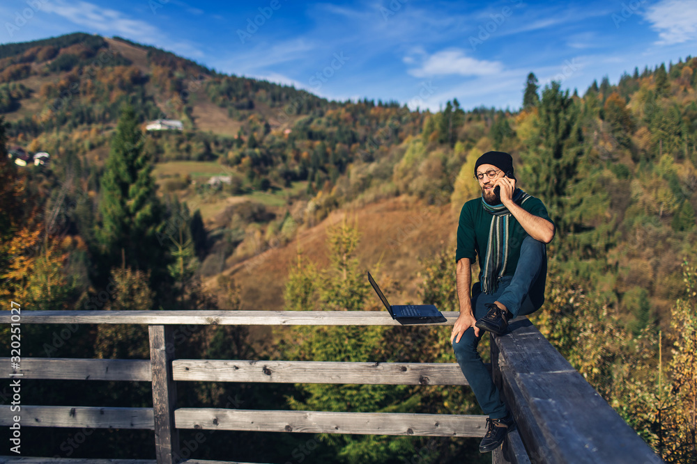 bearded businessman sits on railing, works on laptop and talks on phone. Freelancer hipster on vacation in mountains.