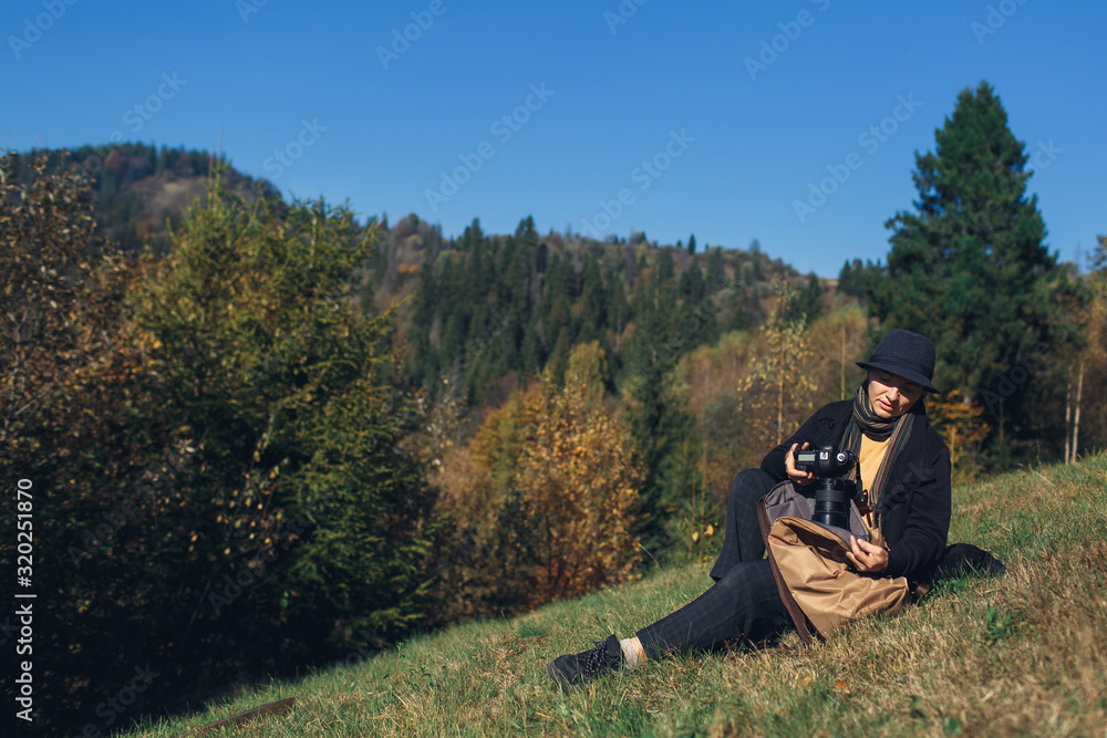 girl photographer sits on slope and takes camera out of backpack in mountains in autumn.