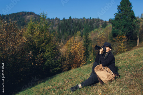 girl photographer with backpack sits on slope and shoot in mountains in autumn.