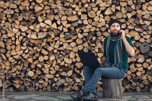 bearded freelancer is working on laptop and talking on phone against background of wall of firewood.