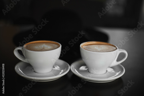 Two white cups of Cappuccino coffee with heart-shaped milk foam. Side view on two cups of latte coffee with heart figure on milk foam on black background. Lifestyle