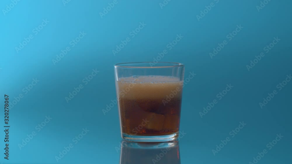 Pouring Cola with ice close up. Cola fizzy drink with ice cubes and bubbles in transparent drinking glass over blue background. Coke Soda closeup. Pouring Cola with ice close up. Cola fizzy drink with ice cubes and bubbles in transparent drinking glass over blue background. Coke Soda closeup.