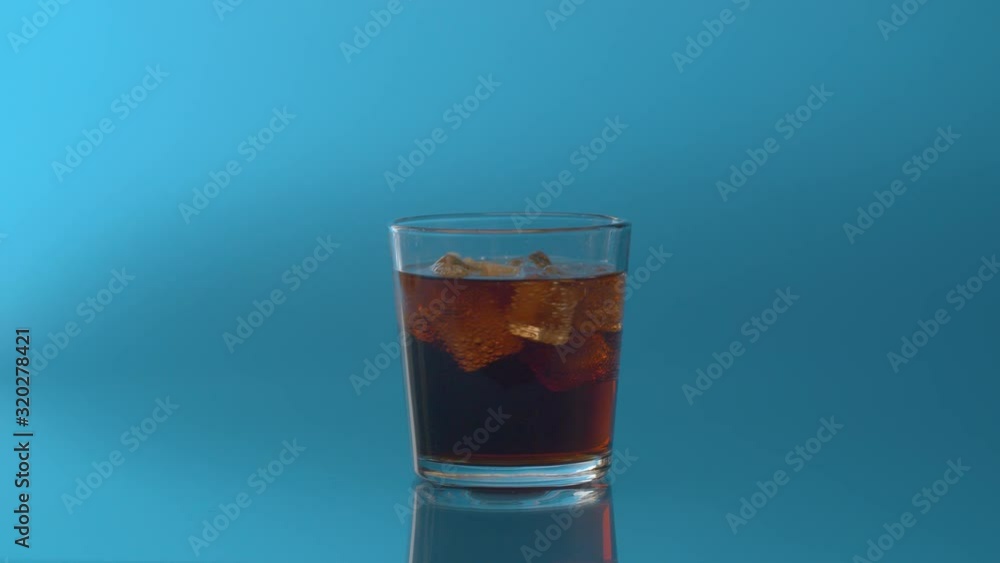 Glass of Cola with Ice and lemon close up. Cola fizzy drink with ice cubes and sliced lemon spinning in transparent drinking glass over blue background. Coke Soda with bubbles closeup. Glass of Cola with Ice and lemon close up. Cola fizzy drink with ice cubes and sliced lemon spinning in transparent drinking glass over blue background. Coke Soda with bubbles closeup.
