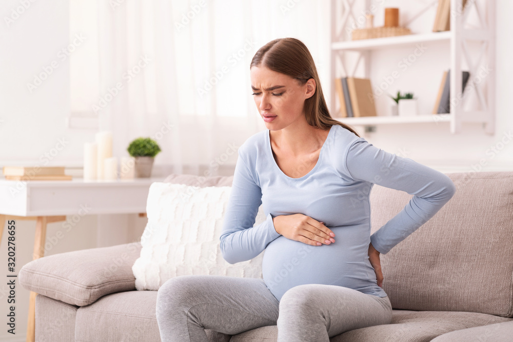 Expectant Woman Having Pain In Lower Back Sitting On Sofa Expectant Woman Having Pain In Lower Back Sitting On Sofa