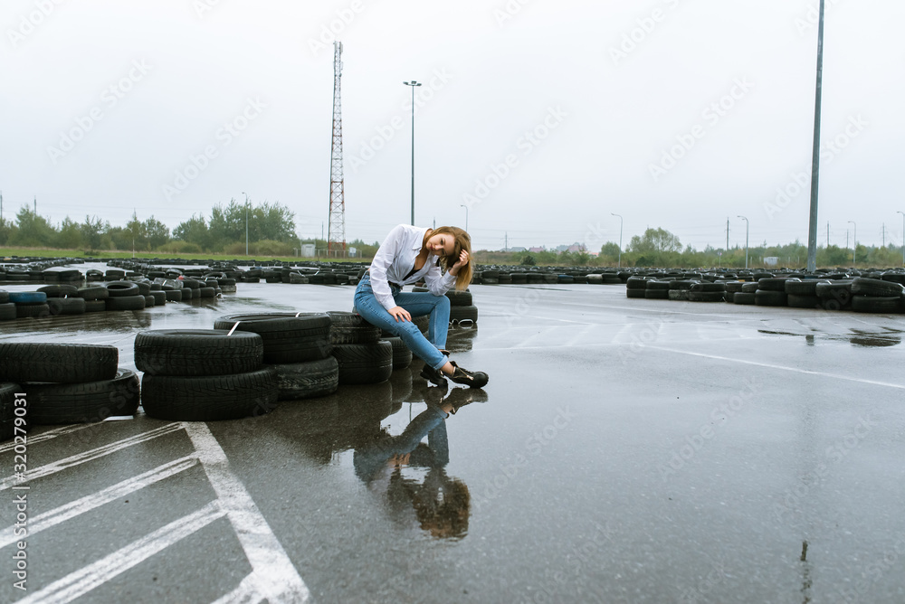 young pretty girl dressed in jeans and white shirt standing during autumn little rain outdoors on wet race track, near old car tires young pretty girl dressed in jeans and white shirt standing during autumn little rain outdoors on wet race track, near old car tires