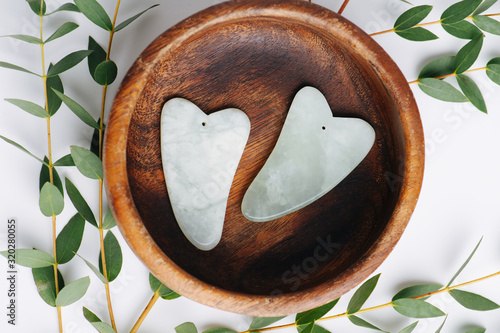 Top view of gua sha jade stones in wooden bowl over branches on white.
