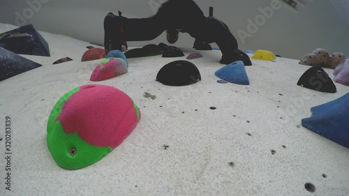 Close up of an indoor climbing gym wall (white) full of different colour  holds seen from below. The wall comes out on an angle. A climber is seen climbing down. photo