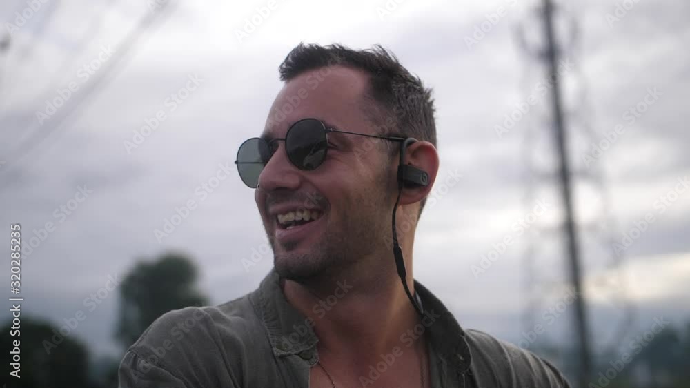 A happy, smiling guy is looking over his shoulder on the side of the road in Bali, Indonesia. Listening to music with his wireless headphones. A happy, smiling guy is looking over his shoulder on the side of the road in Bali, Indonesia. Listening to music with his wireless headphones.