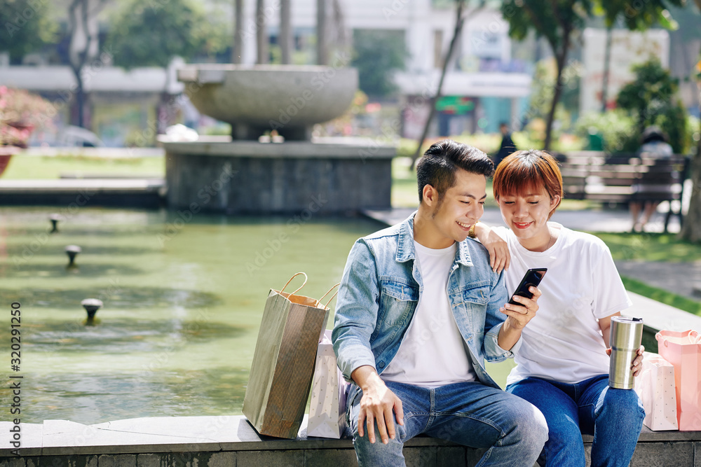 Horizontal shot of young man and woman in love sitting together with shopping bags against fountains watching something on smartphone