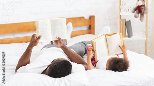 Valokuva Black dad and little daughter reading books, laying on bed