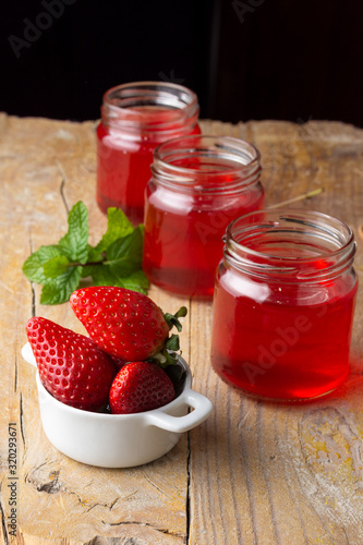 Top view of bowl of strawberries, three recycled glass jars with strawberry jelly and mint branch, on rustic wooden table, in vertical, with dark background in vertical