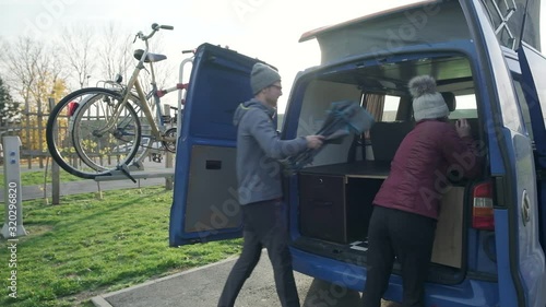 A young couple packing up a chair and frisbee in the back of a blue campervan. On holiday on a sunny day playing in the field. A couple travelling in a campervan. photo