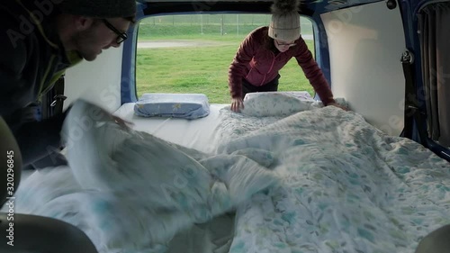 A man and women making a bed in the back of a campervan. The camera set up looking into the back of the van with the back doors open. A couple travelling in a campervan. photo