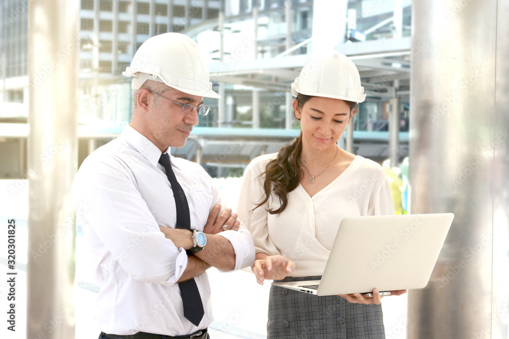 Two engineer wearing white safety helmet using a laptop together, male and female discussing about project work while walking on the way outside office in downtown city Two engineer wearing white safety helmet using a laptop together, male and female discussing about project work while walking on the way outside office in downtown city