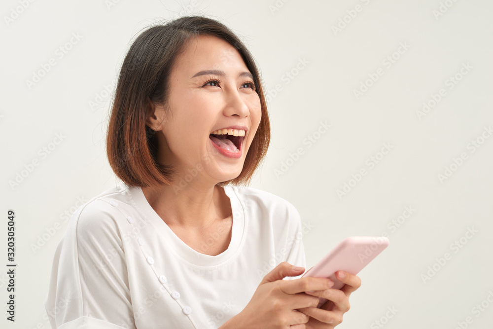 Cheerful asian woman standing isolated over white background, holding mobile phone, celebrating