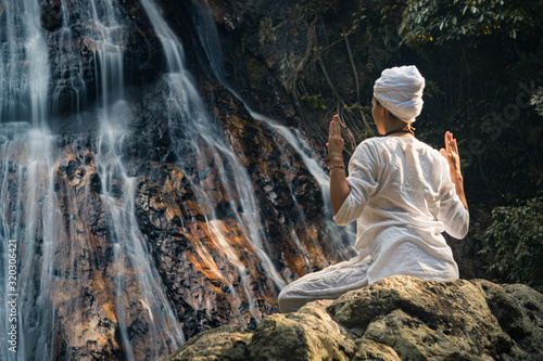 Young adult woman in white clothes sits on a rock against the background of a waterfall and practices yoga