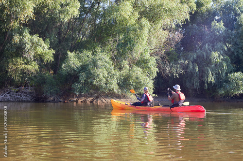 Man and a woman paddle kayak on a river near the shore overgrown with willows