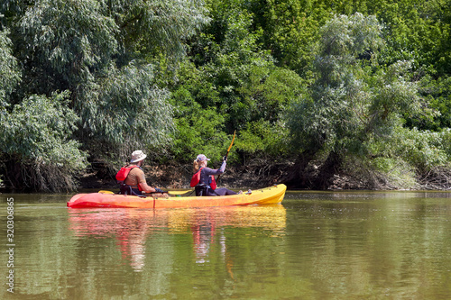 Man and a woman paddle kayak on a river near the shore overgrown with willows