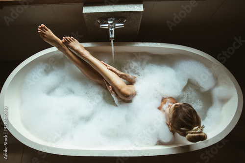 Woman relaxing in foam bath with bubbles in dark bathroom by window Fototapeta