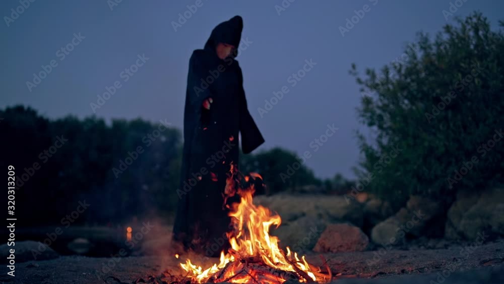 Witch evil in black gown near fire at night. Mysterious wizard doing shamanism over the flame. Scary figure in black making ritual. Halloween night. Witch evil in black gown near fire at night. Mysterious wizard doing shamanism over the flame. Scary figure in black making ritual. Halloween night.