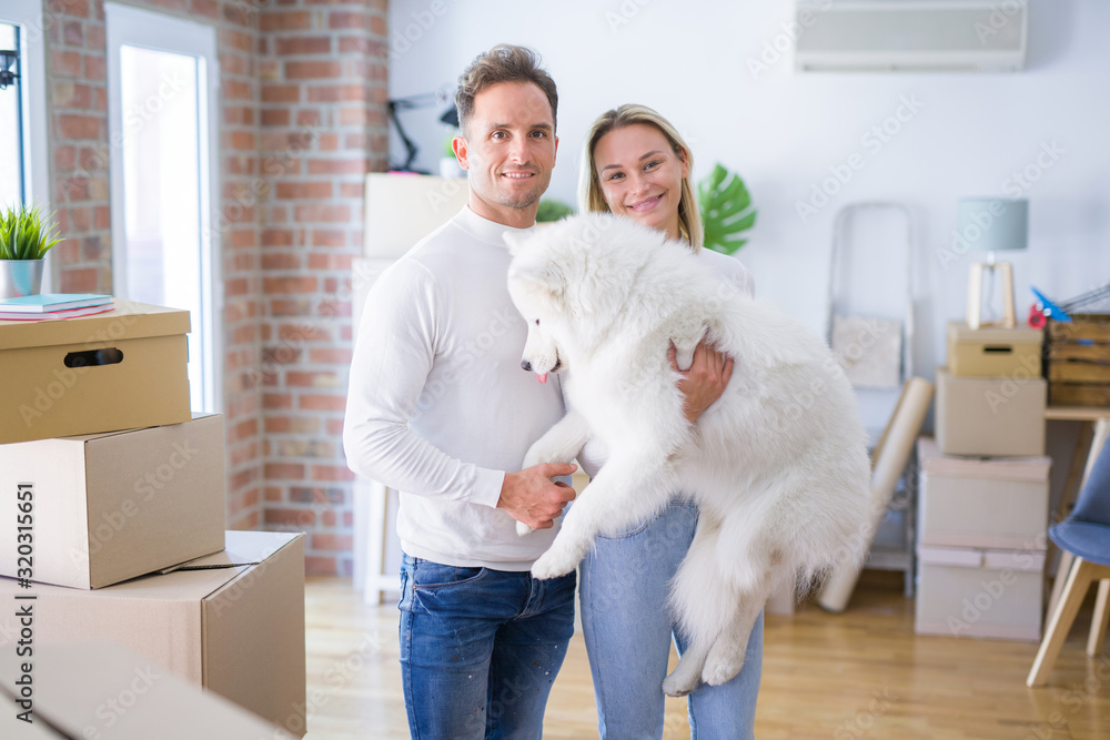 Young beautiful couple with dog standing at new home around cardboard boxes Young beautiful couple with dog standing at new home around cardboard boxes