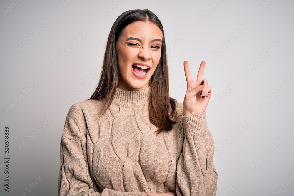Young beautiful brunette woman wearing casual sweater standing over white background smiling with happy face winking at the camera doing victory sign. Number two. Young beautiful brunette woman wearing casual sweater standing over white background smiling with happy face winking at the camera doing victory sign. Number two.