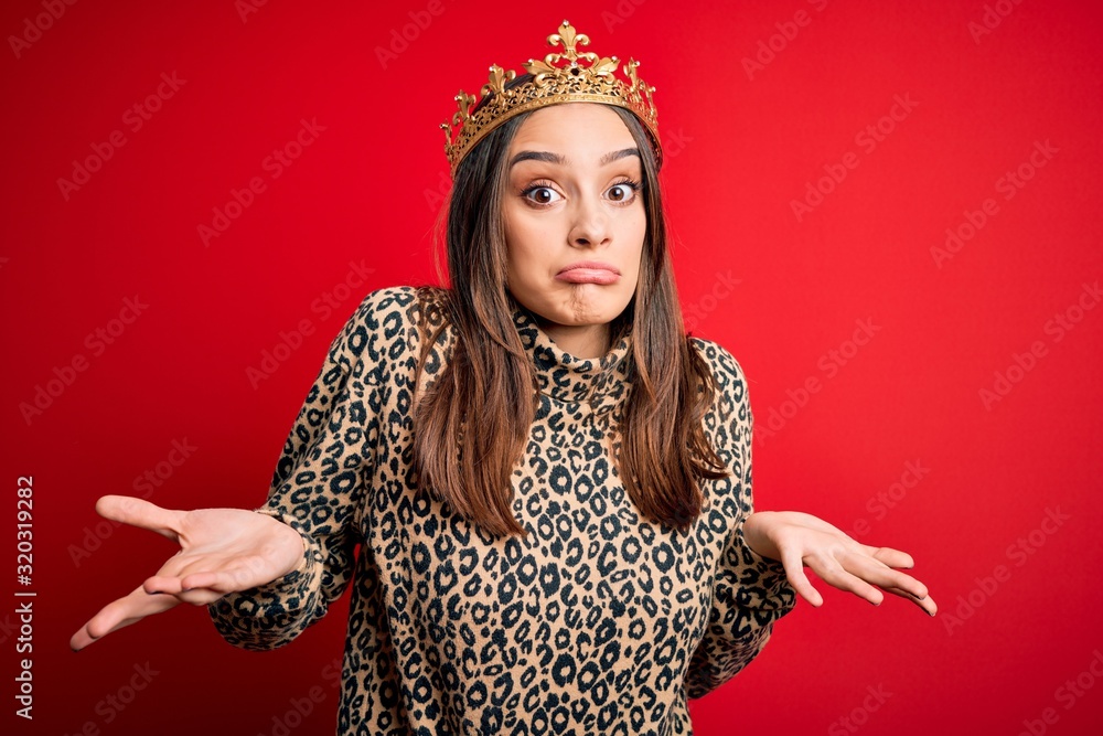 Young beautiful brunette woman wearing golden crown queen over isolated red background clueless and confused expression with arms and hands raised. Doubt concept.
