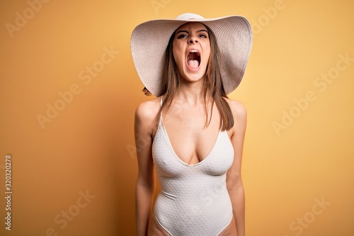Young beautiful brunette woman on vacation wearing swimsuit and summer hat angry and mad screaming frustrated and furious, shouting with anger. Rage and aggressive concept.