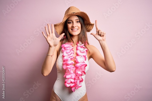 Young beautiful brunette woman on vacation wearing swimsuit and Hawaiian flowers lei showing and pointing up with fingers number seven while smiling confident and happy.