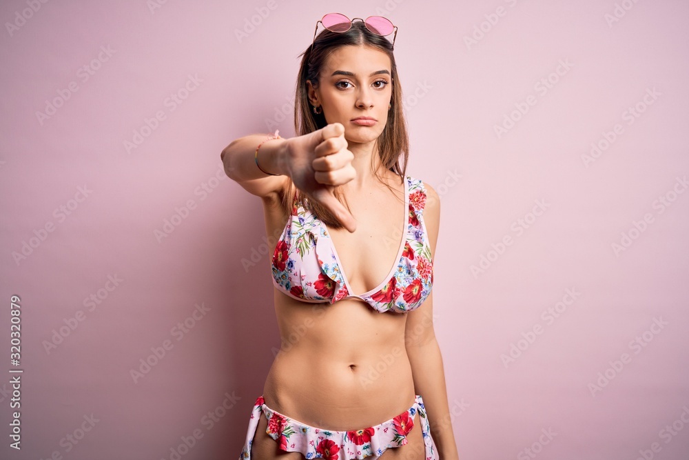 Naklejka premium Young beautiful woman on vacation wearing bikini and sunglasses over pink background looking unhappy and angry showing rejection and negative with thumbs down gesture. Bad expression.