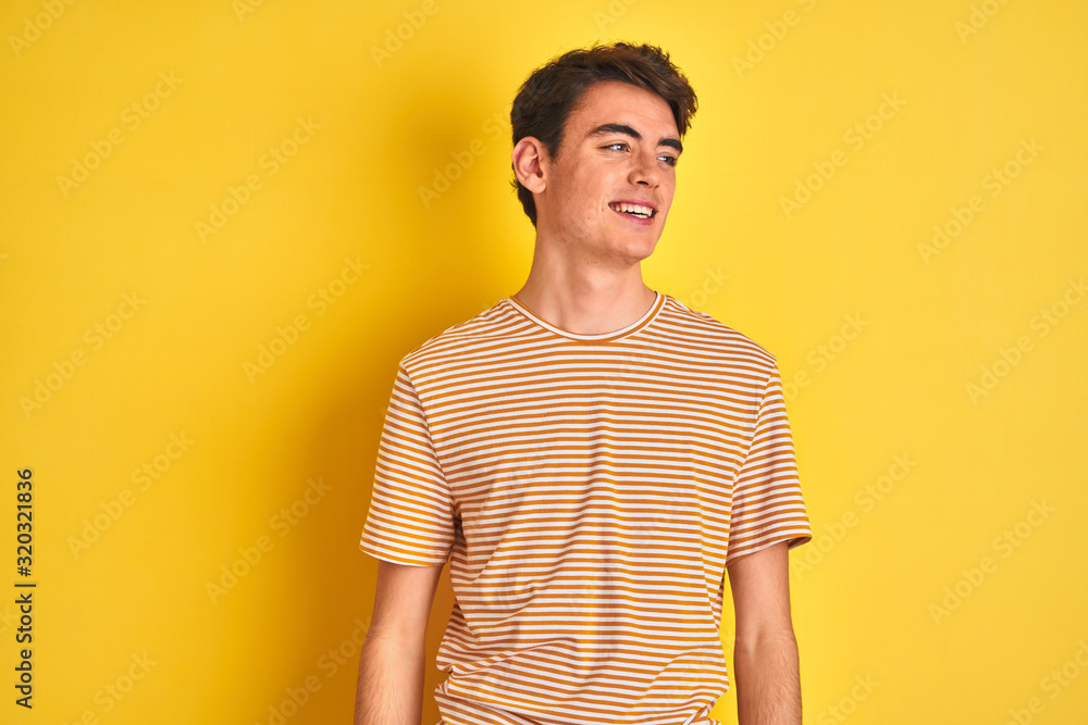 Teenager boy wearing yellow t-shirt over isolated background looking away to side with smile on face, natural expression. Laughing confident.