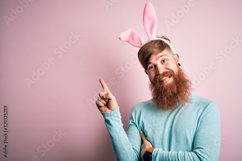 Hipster Irish man with beard wearing easter rabbit ears over isolated pink background with a big smile on face, pointing with hand and finger to the side looking at the camera. photo