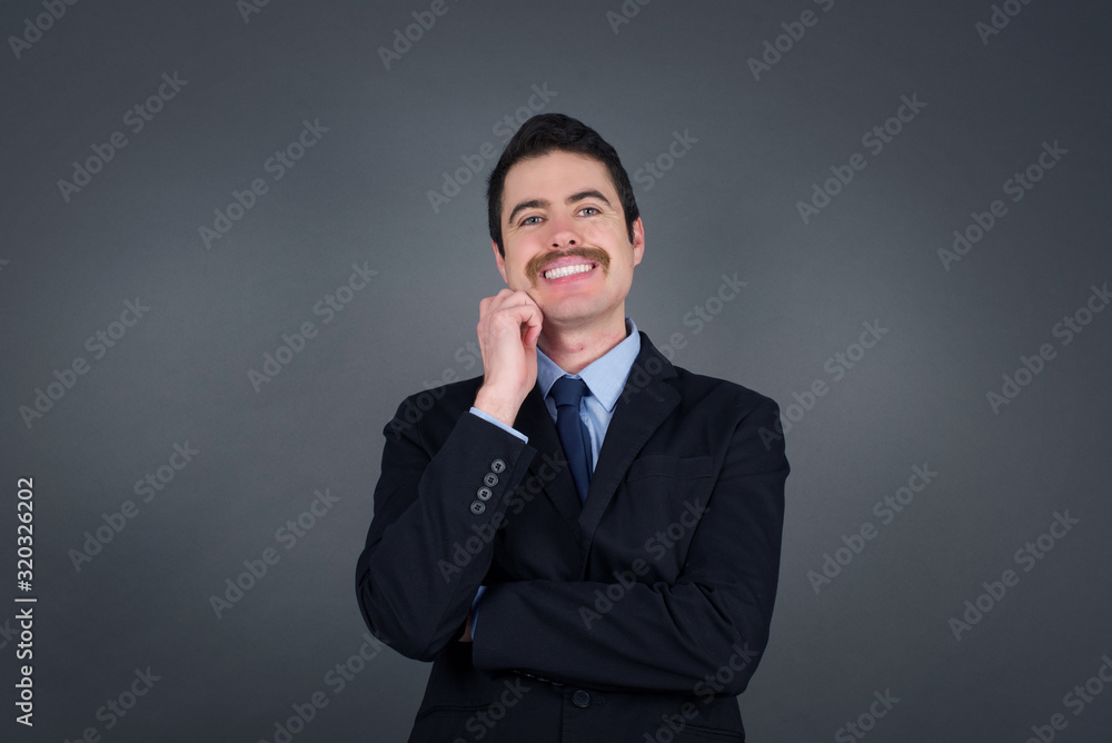 Image of cheerful pretty young caucasian man standing against gray wall with hand near face. Looking with glad expression at the camera after listening to good news. Confident man. Image of cheerful pretty young caucasian man standing against gray wall with hand near face. Looking with glad expression at the camera after listening to good news. Confident man.