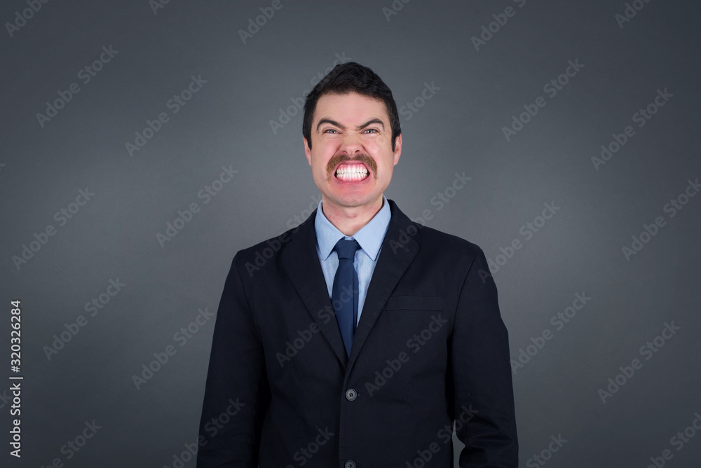 Mad crazy man clenches teeth angrily, being annoyed with coming noise, dressed in fashionable clothes, isolated over gray background. Negative feeling concept. Mad crazy man clenches teeth angrily, being annoyed with coming noise, dressed in fashionable clothes, isolated over gray background. Negative feeling concept.