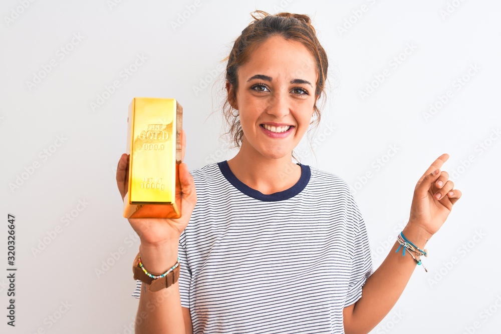 Young beautiful redhead woman holding gold ingot over isolated background very happy pointing with hand and finger to the side Young beautiful redhead woman holding gold ingot over isolated background very happy pointing with hand and finger to the side