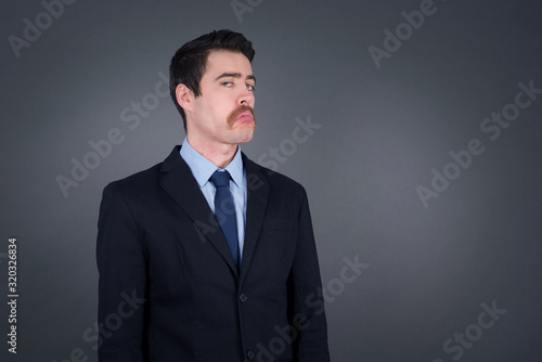 Beautiful businessman with snobbish expression curving lips and raising eyebrows, looking with doubtful and skeptical expression, suspect and doubt. Standing indoors over gray background. © Jihan