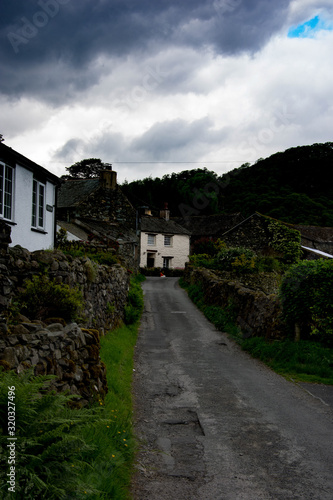 A small cottage and other farm buildings are positioned at the end of a narrow country lane with dark stormy clouds imposing above. 