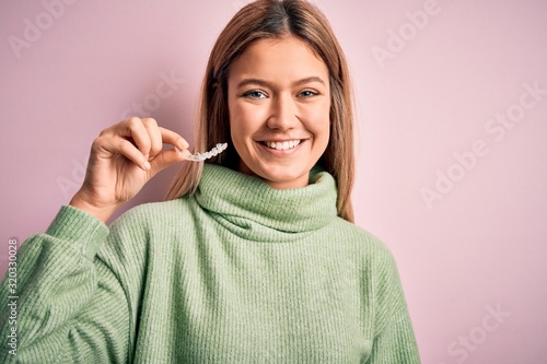 Young beautiful woman holding aligner standing over isolated pink background with a happy face standing and smiling with a confident smile showing teeth photo