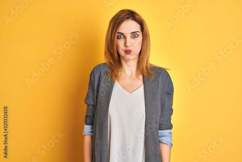 Redhead caucasian business woman over yellow isolated background puffing cheeks with funny face. Mouth inflated with air, crazy expression. © Krakenimages.com