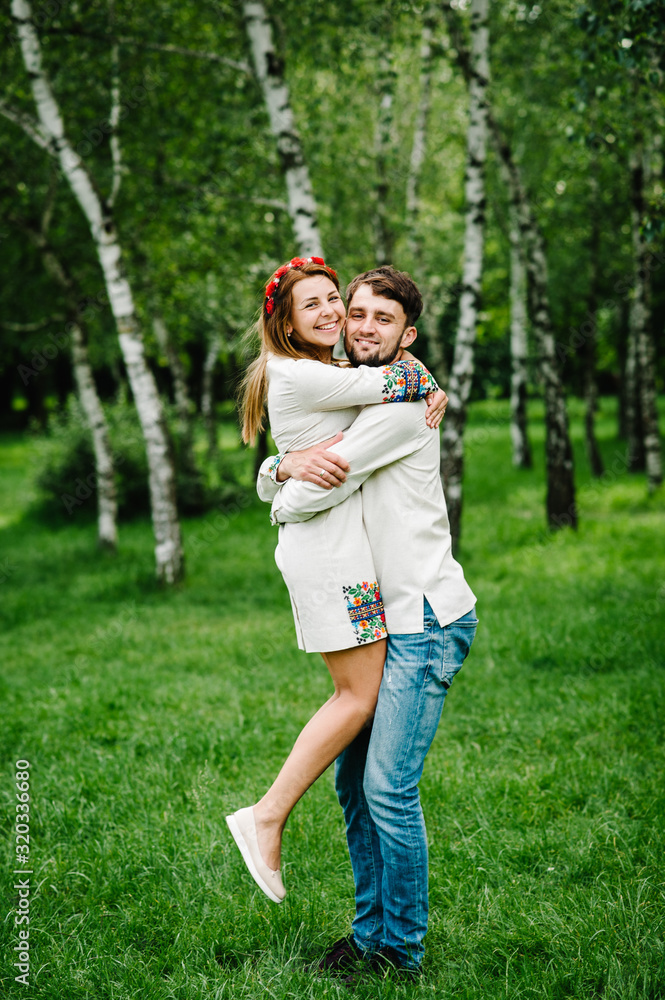 Fototapeta premium The man raised the woman to her arms. Happy family on the background of birches on nature. Close up. Looking at the camera. Full length. Embroidered shirt and handmade dress.