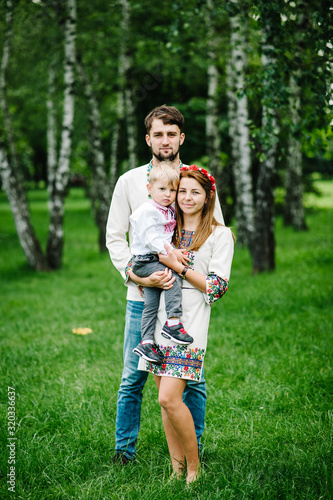 Mother, Dad hold a son on the arms in embroidery. Happy family on the background of birches on nature. Close up. Looking at the camera. Full length. Embroidered shirt and handmade dress.