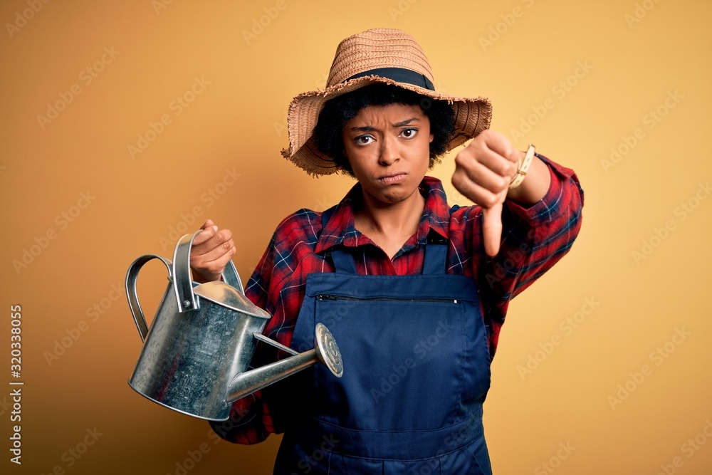 Young African American farmer woman with curly hair wearing apron and hat using water can with angry face, negative sign showing dislike with thumbs down, rejection concept Young African American farmer woman with curly hair wearing apron and hat using water can with angry face, negative sign showing dislike with thumbs down, rejection concept