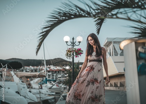 Stylish young woman with brown eyes, straight dark hair and a lovely smile, dressed in a long expensive dress, walks along the pier next to the yacht club and the sea, yachts and boats