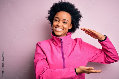 Young African American afro sportswoman with curly hair wearing sportswear doin sport gesturing with hands showing big and large size sign, measure symbol. Smiling looking at the camera. Measuring