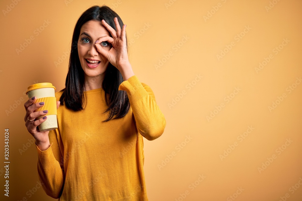 Young woman with blue eyes holding cup of coffee standing over yellow background with happy face smiling doing ok sign with hand on eye looking through fingers Young woman with blue eyes holding cup of coffee standing over yellow background with happy face smiling doing ok sign with hand on eye looking through fingers