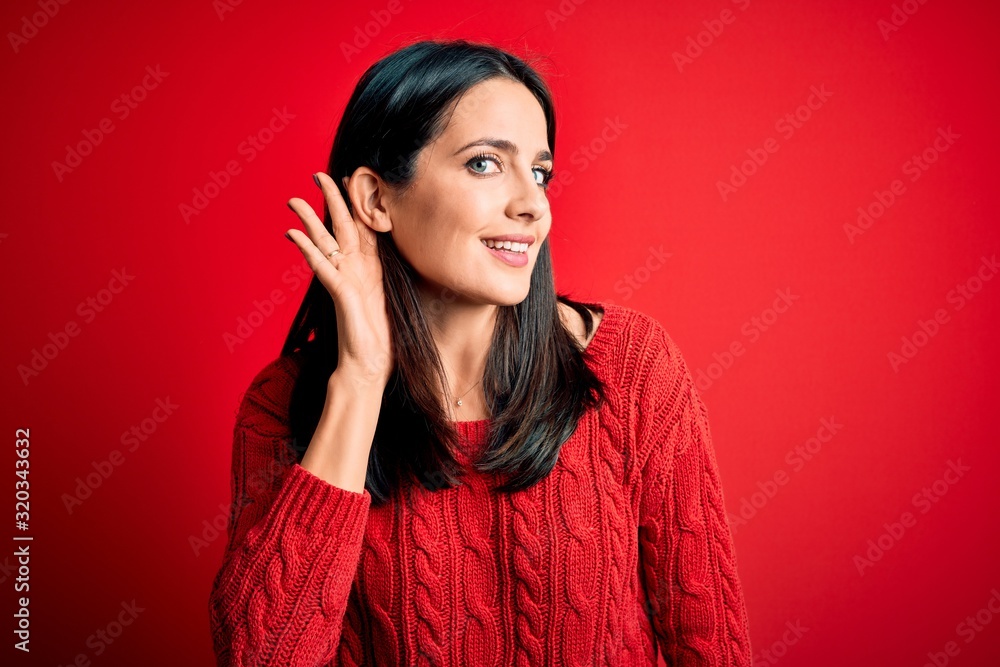 Young brunette woman with blue eyes wearing casual sweater over isolated red background smiling with hand over ear listening an hearing to rumor or gossip. Deafness concept. Young brunette woman with blue eyes wearing casual sweater over isolated red background smiling with hand over ear listening an hearing to rumor or gossip. Deafness concept.